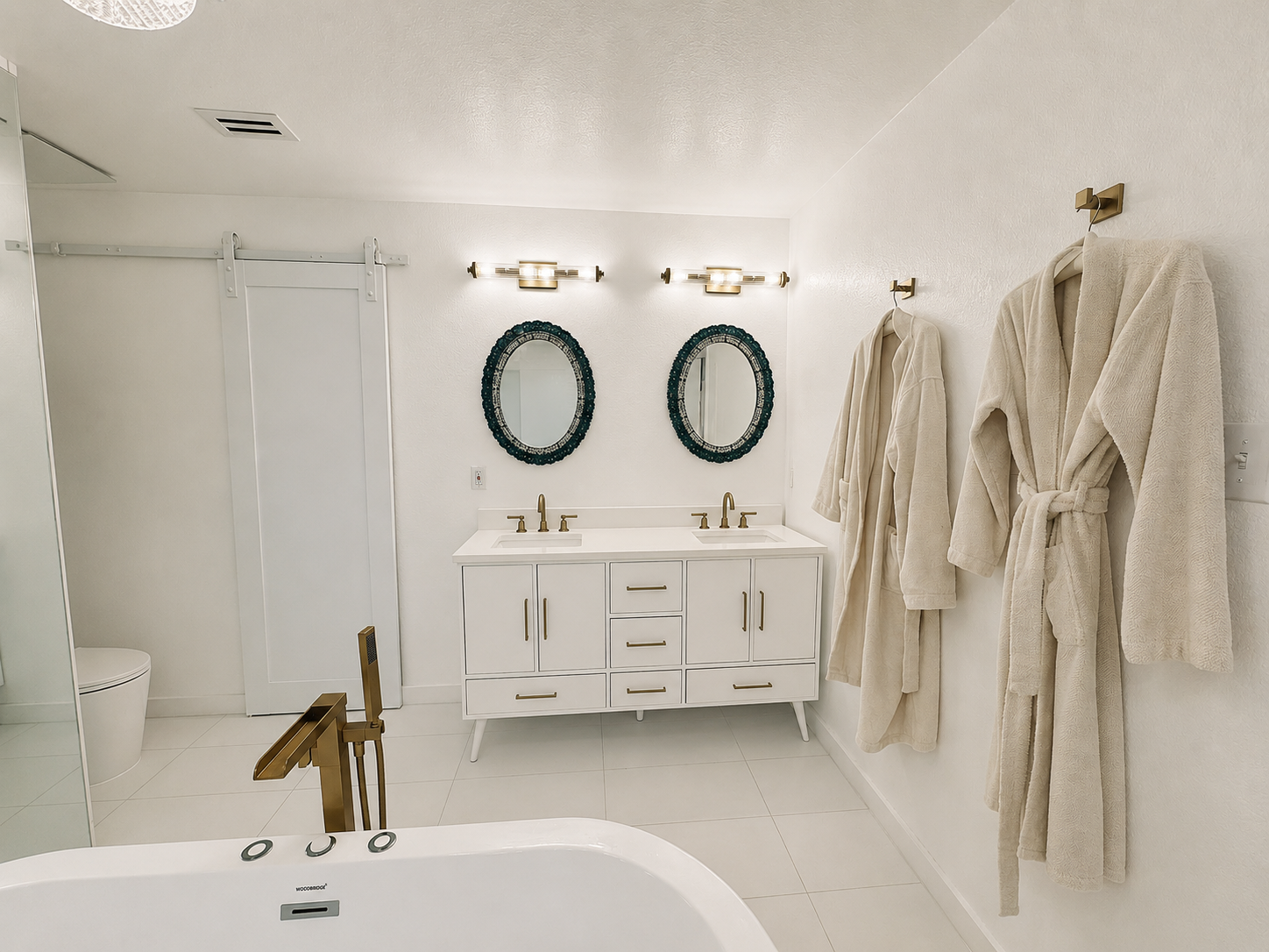 Bright white bathroom with freestanding tub, brass fixtures, double vanity, oval mirrors, and hanging robes