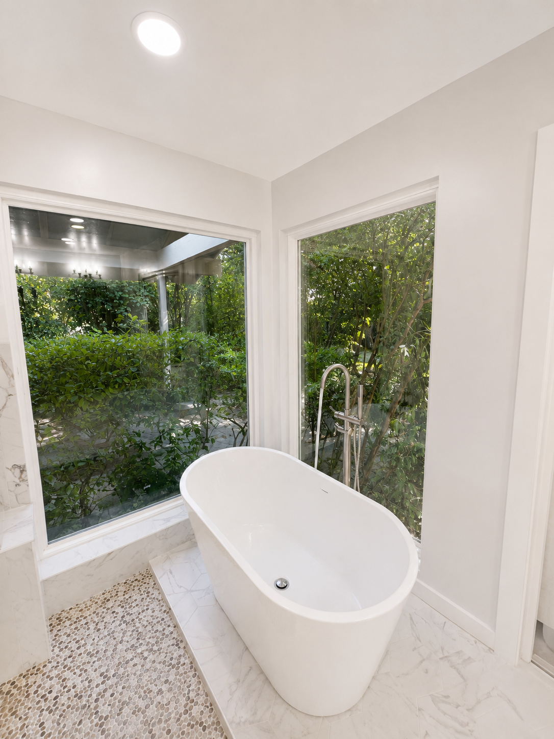 Modern bathroom with a freestanding white tub beside large corner windows overlooking greenery