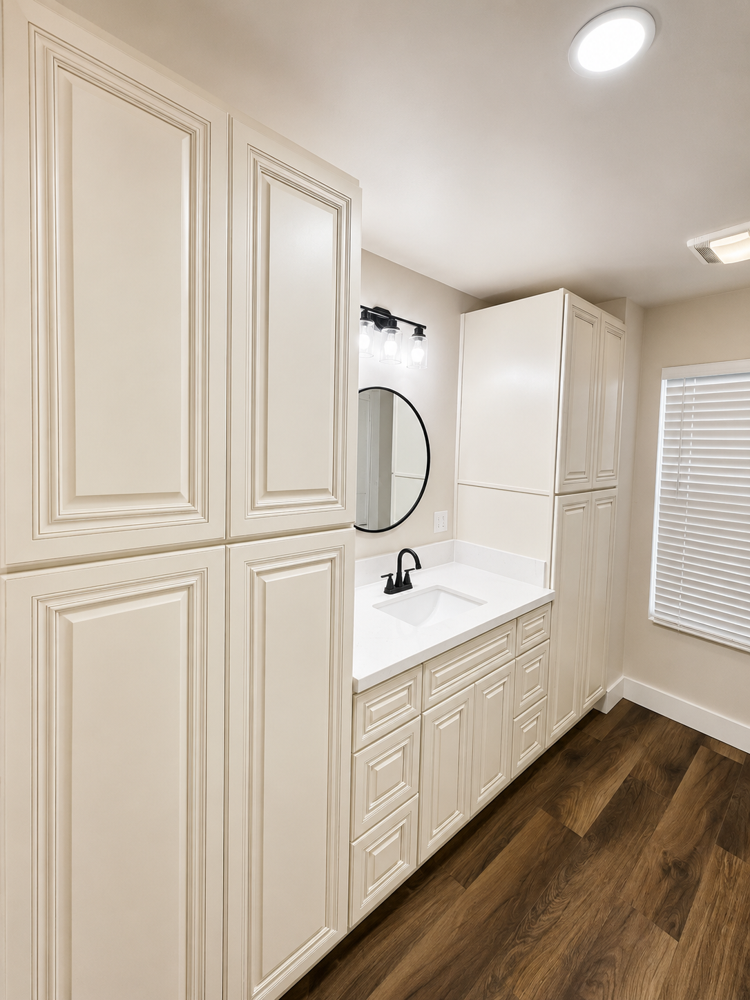 Bright bathroom with tall white cabinets, vanity sink, round mirror, and wood-look floor.