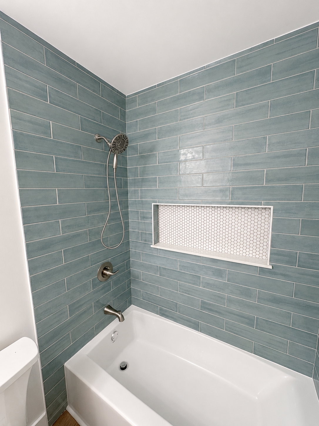 Light blue tiled bathtub alcove with shower fixtures and a small white vent panel on the wall