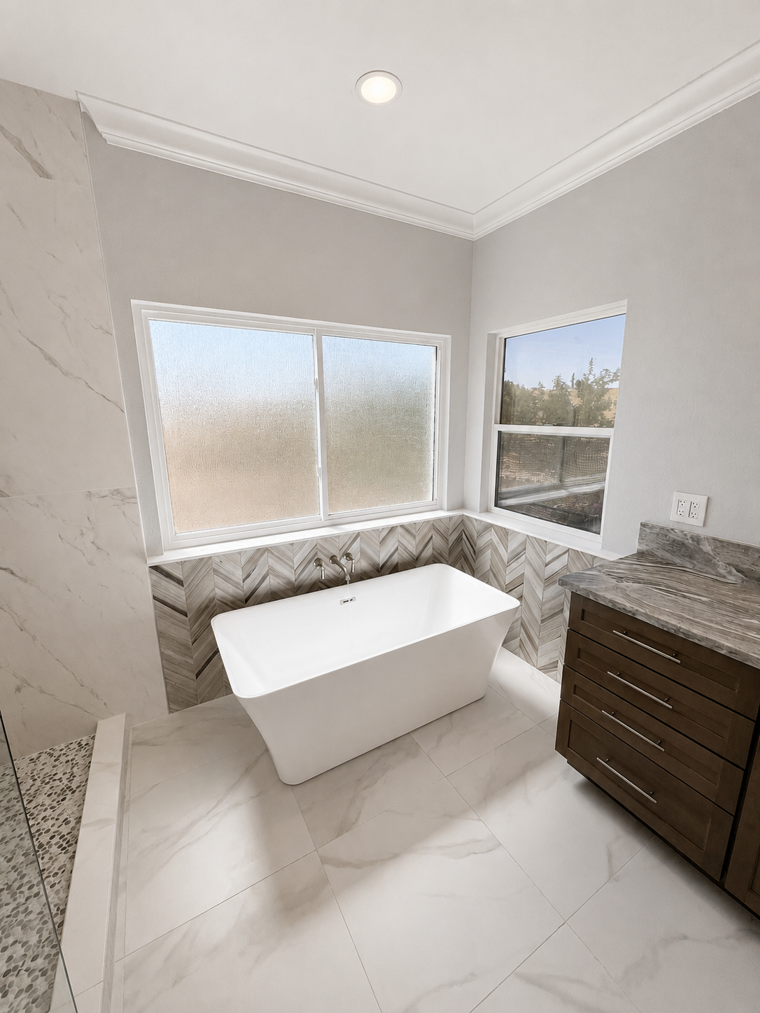 Modern bathroom with freestanding tub, gray tile, frosted windows, and dark wood vanity.