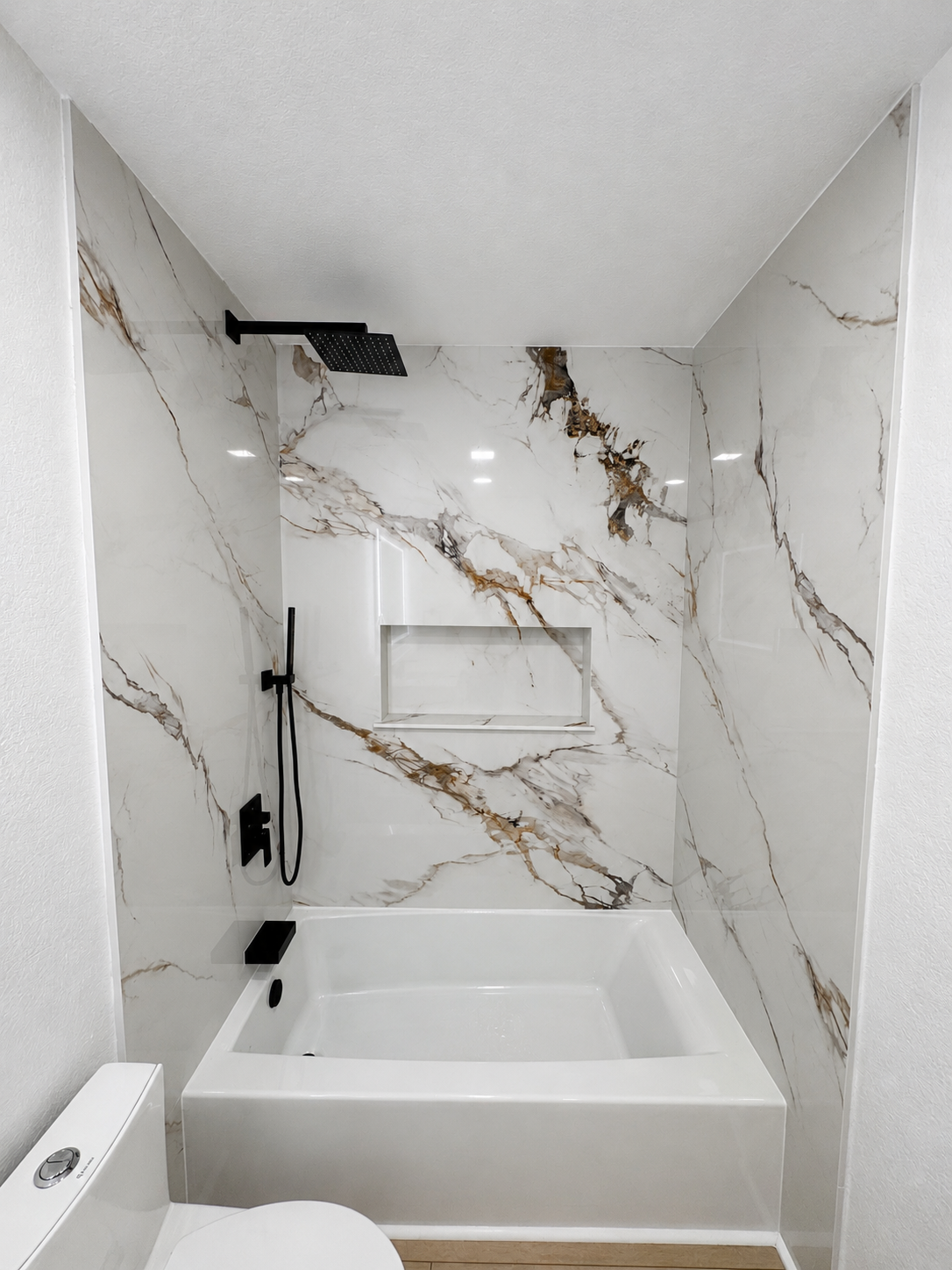 Modern white tiled shower with gold veining, black fixtures, and a white bathtub in a small bathroom