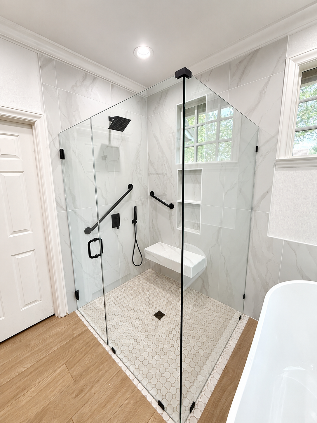 Glass-enclosed walk-in shower with white tile, black fixtures, and wood-look floor in a bright bathroom