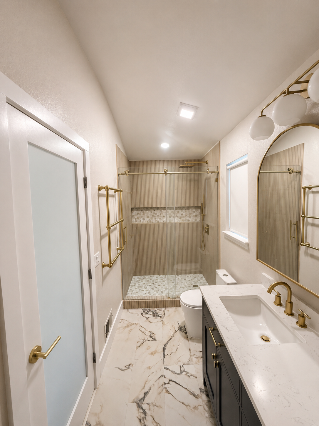 Bright bathroom with marble vanity, glass shower, brass fixtures, and frosted window.