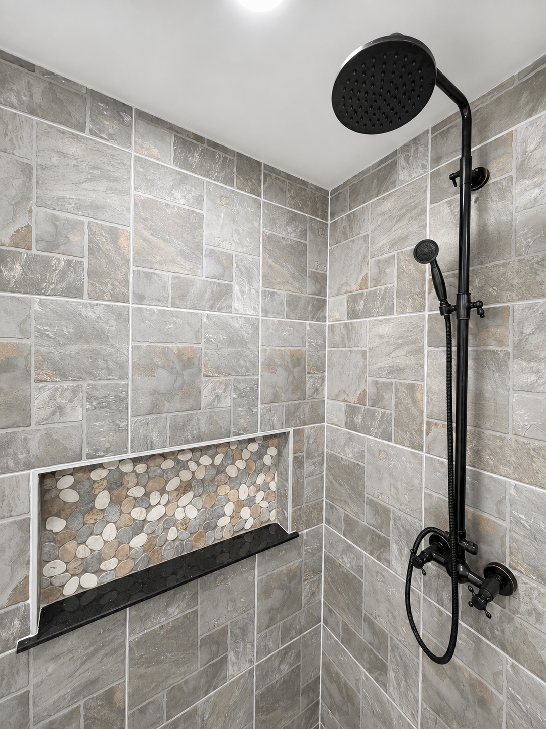 Corner walk-in shower with gray tile, pebble accent shelf, and matte black rainfall showerhead and fixtures