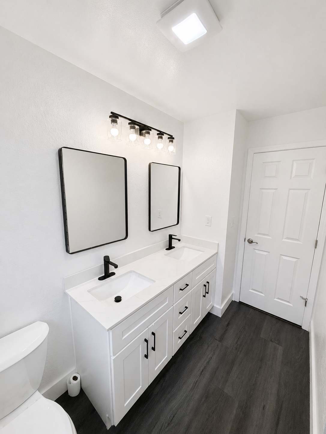 Bright white bathroom with double vanity, two mirrors, black fixtures, and dark wood flooring.