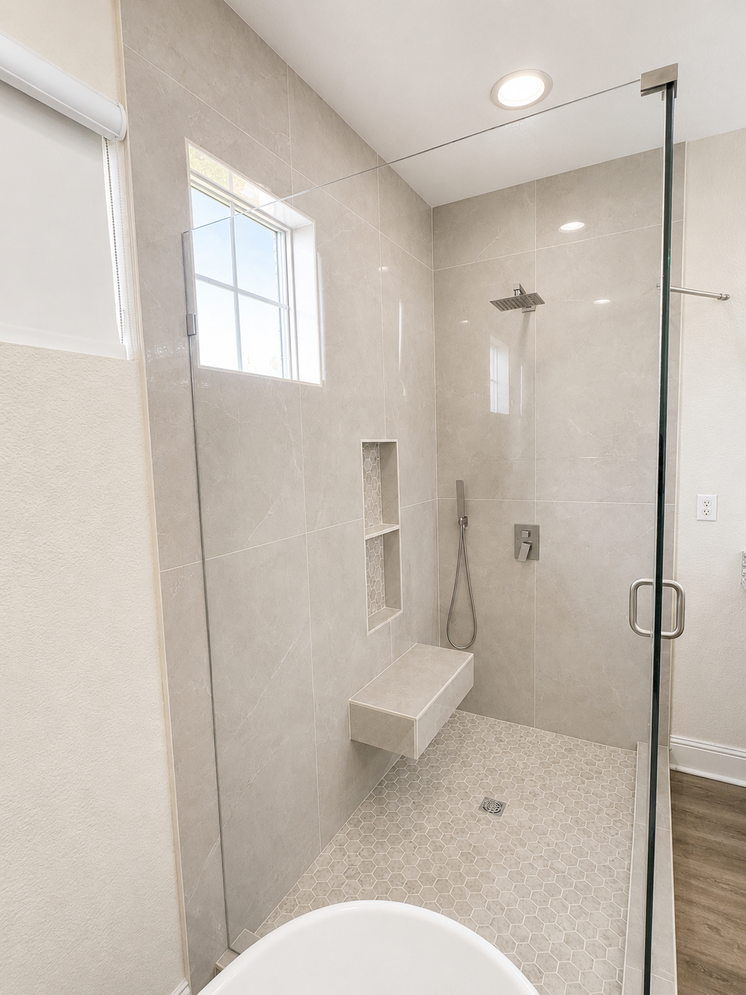 Accessible walk-in shower with bench, grab bars, shower niche, and glass door in a light tiled bathroom