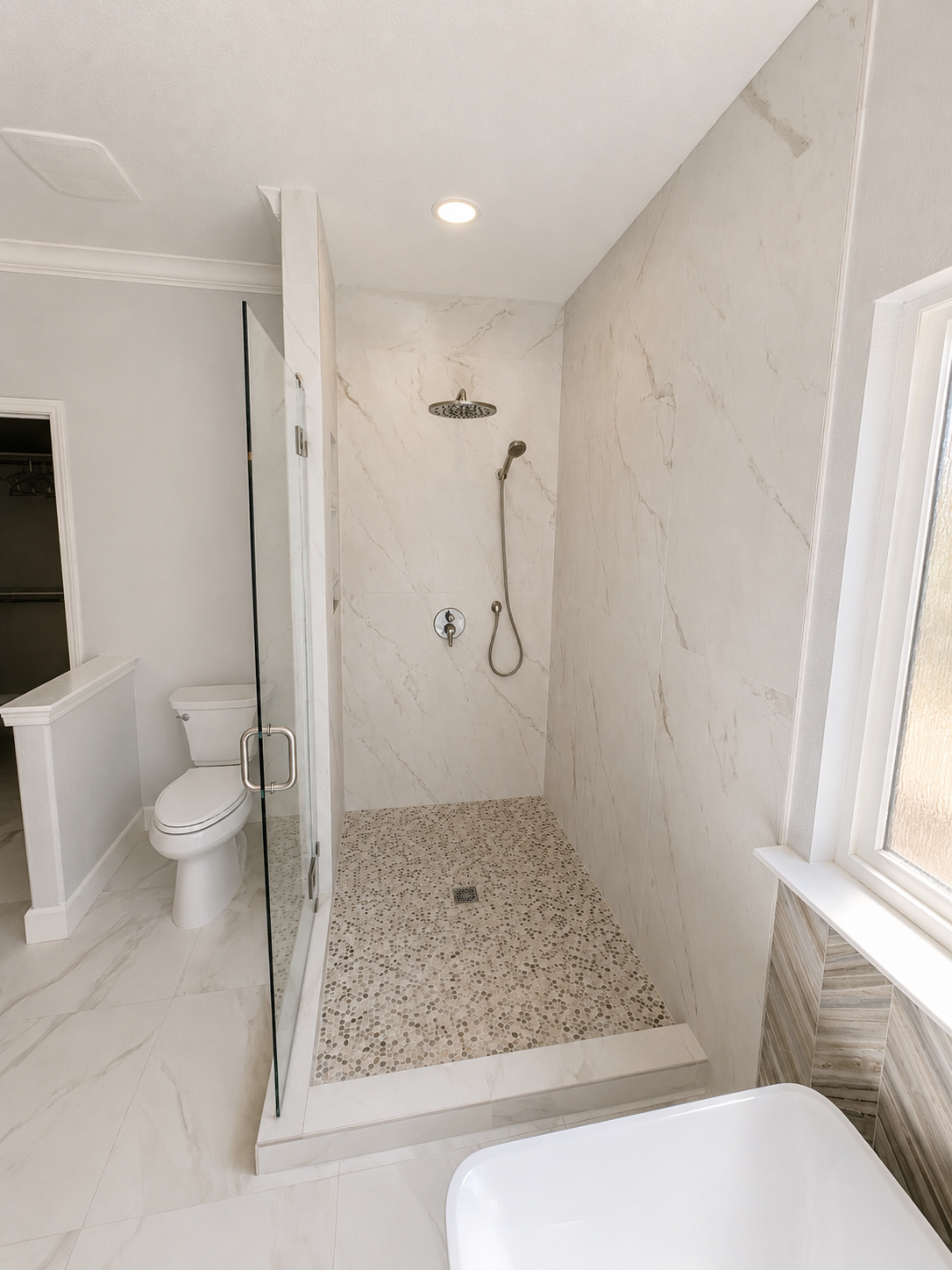 Small white bathroom with a walk-in tiled shower, toilet, and window beside the shower.