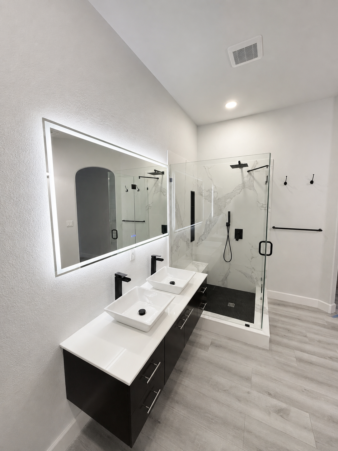 Bright modern bathroom with double vanity, lit mirror, glass shower, and white walls and floors