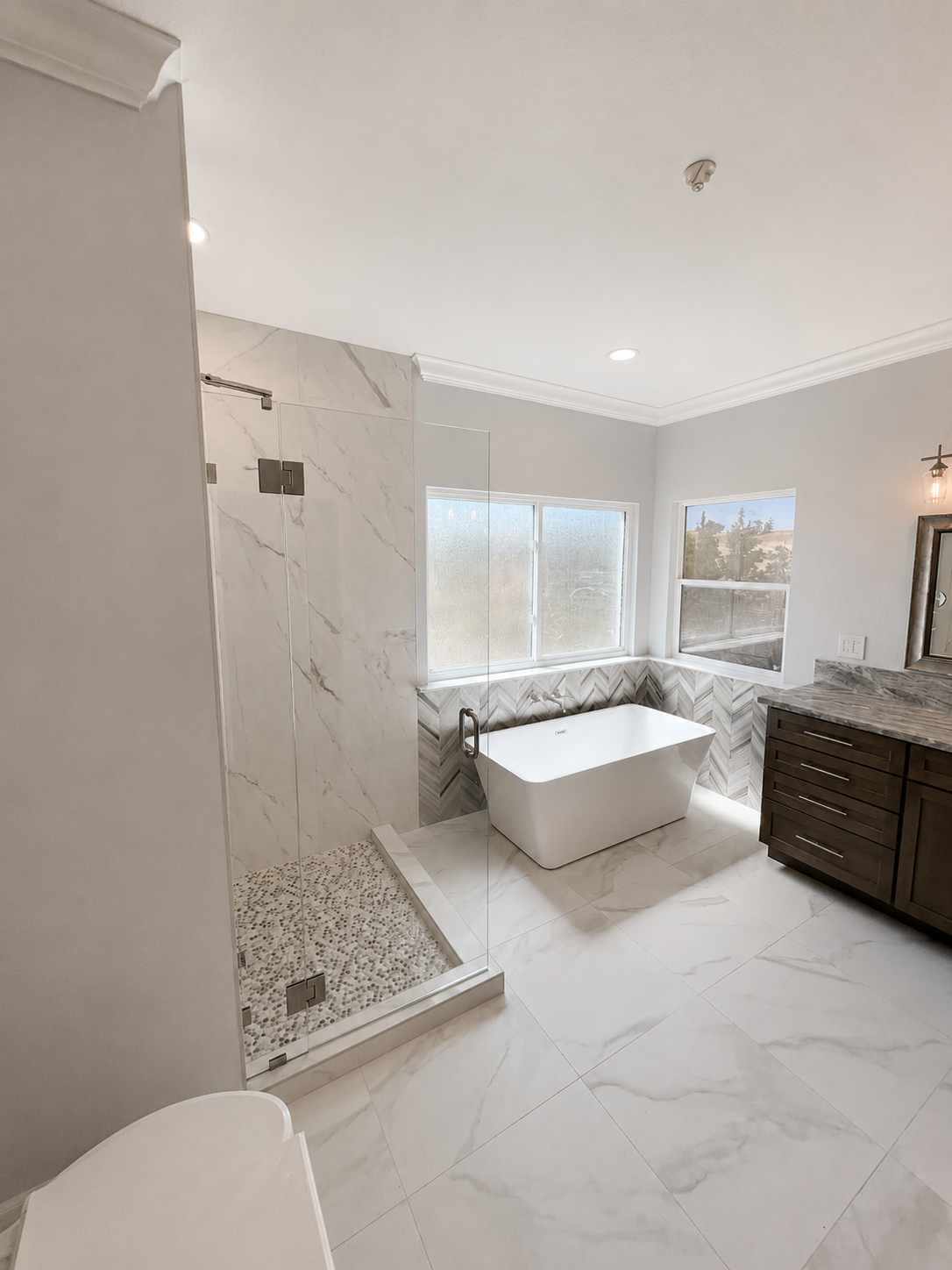 Bright bathroom with marble tile shower, freestanding tub by windows, and dark vanity cabinet.