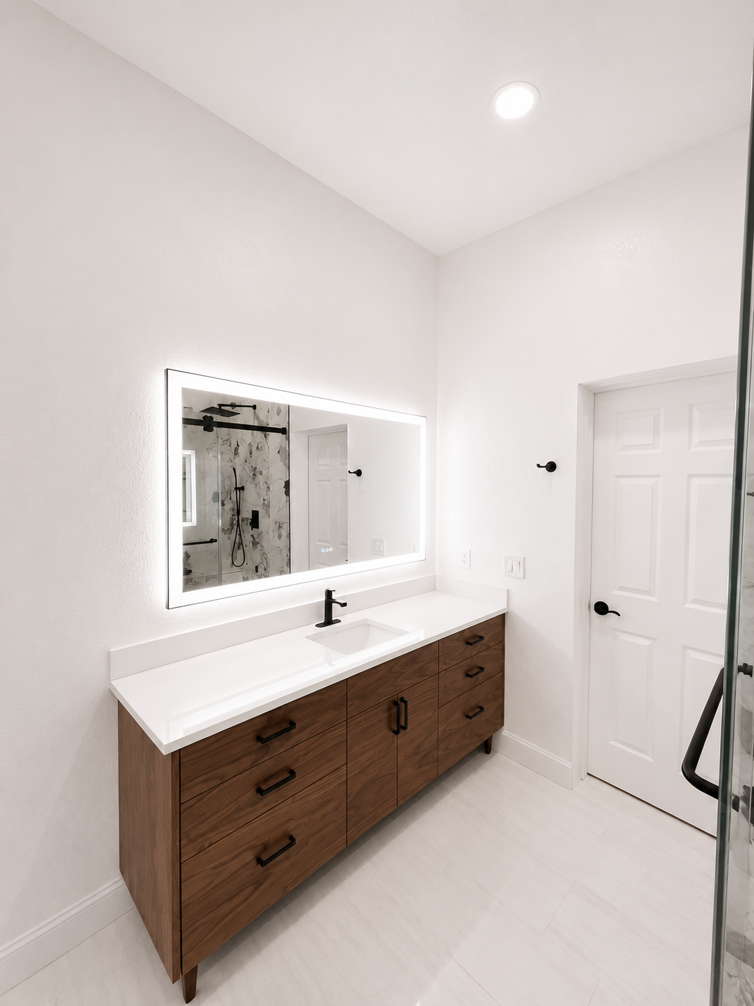 Bright white bathroom with wooden vanity, illuminated mirror, sink, and shower visible in reflection.
