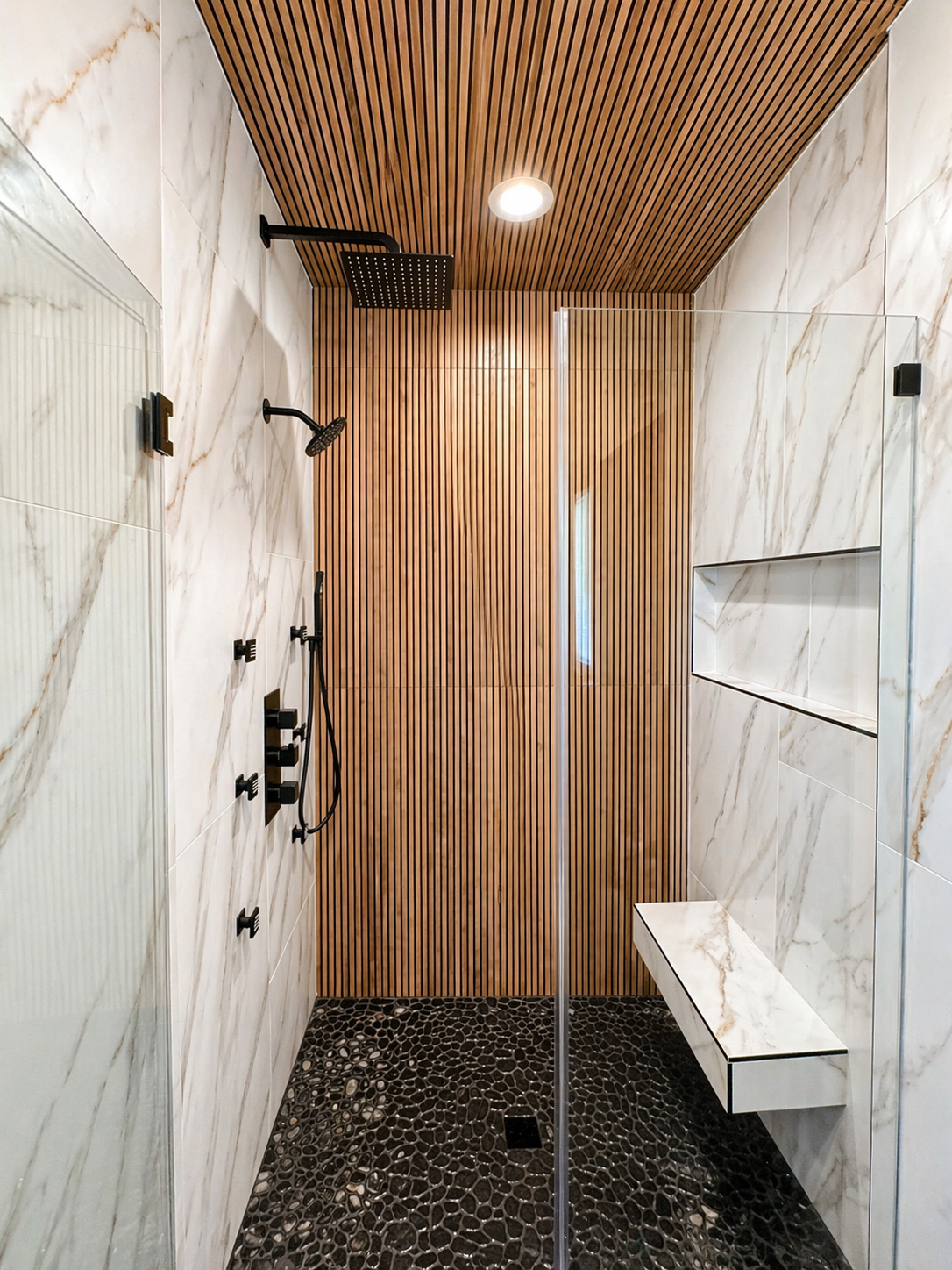 Modern shower with marble walls, wood slat ceiling, black fixtures, and pebble floor.