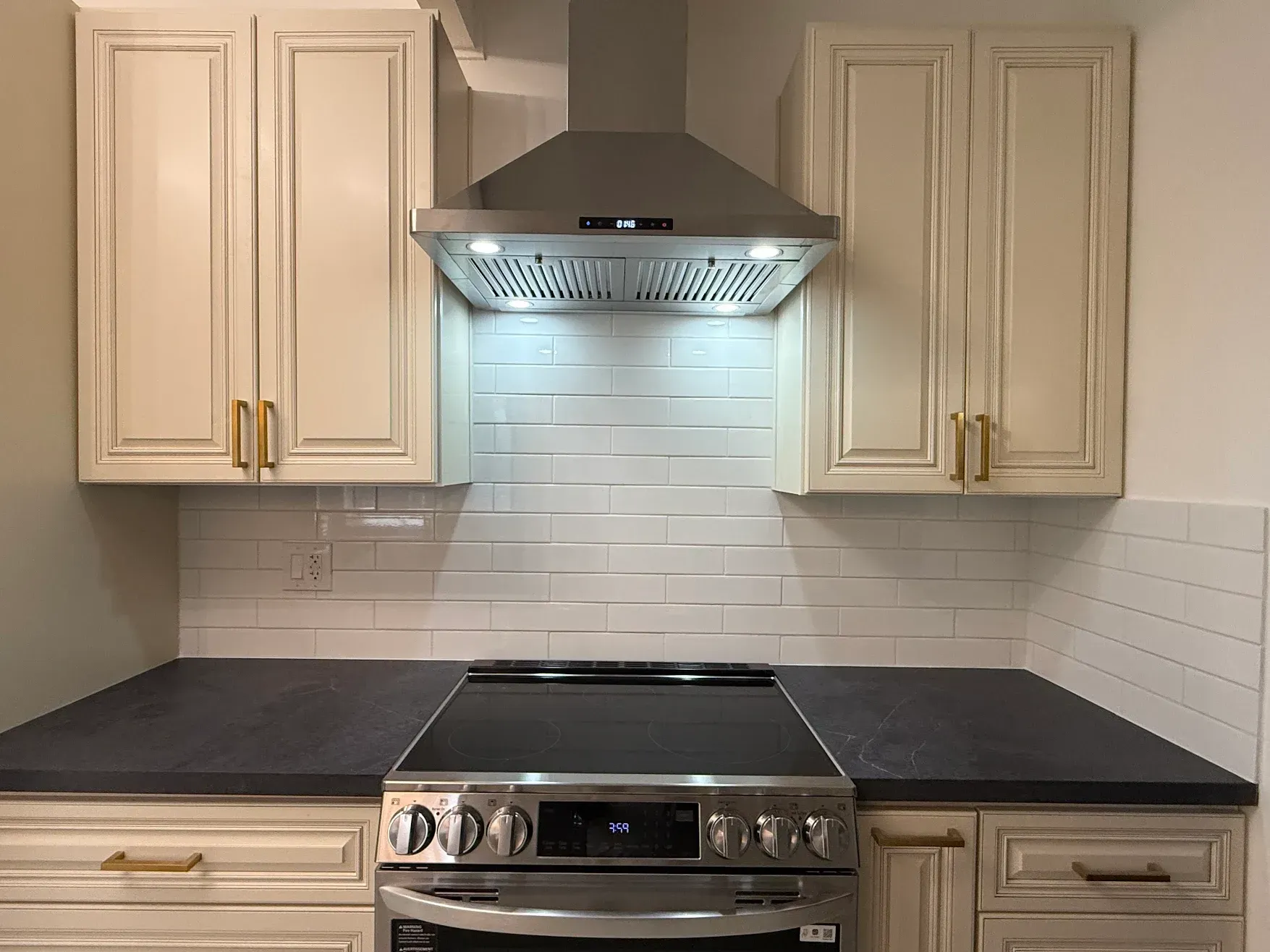 Modern kitchen with white cabinets, black countertops, stainless range, and lit hood over white tile backsplash