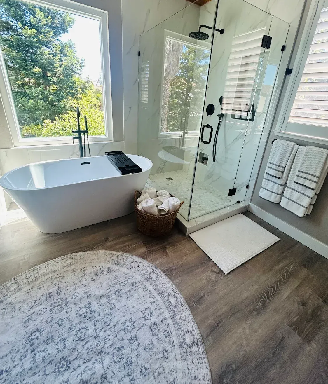 Bright modern bathroom with freestanding tub, glass shower, window, and woven basket on wood floor