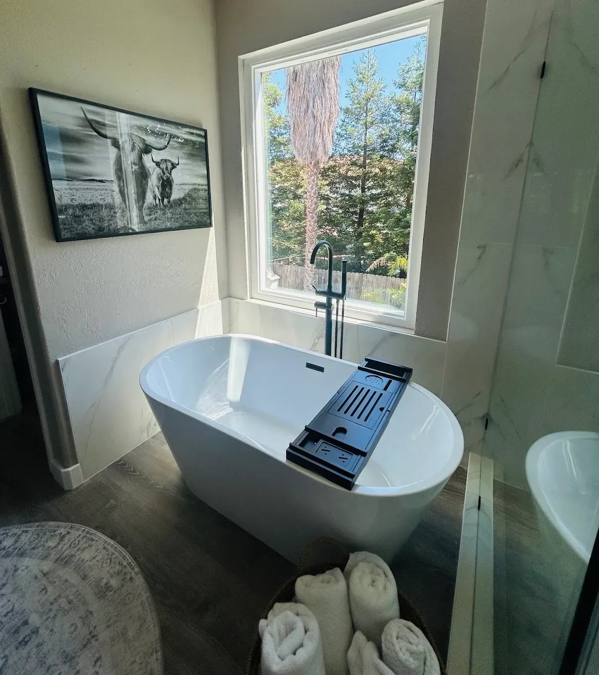 Modern bathroom with a freestanding tub, large window, wall art, and rolled towels on the floor