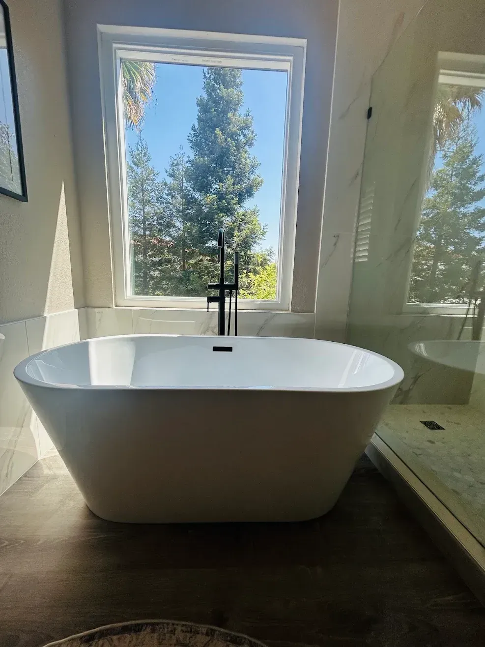 Modern bathroom with a freestanding white tub, black faucet, and large windows overlooking trees.