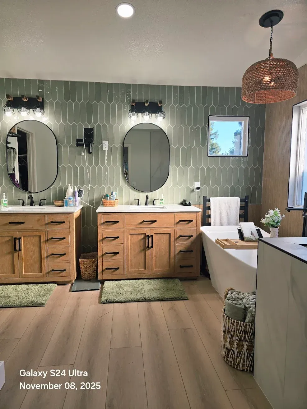 Bathroom with double wood vanity, oval mirrors, tiled walls, and freestanding tub beside a window.