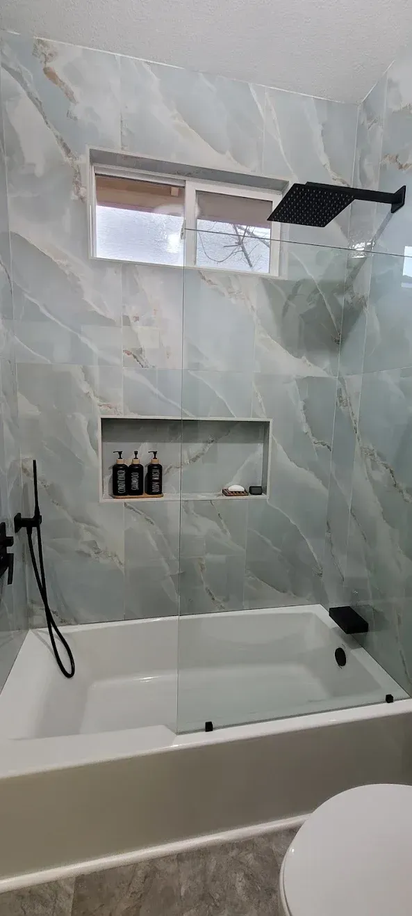 Modern bathroom with gray tiled walls, a white tub, and a small window above the shower niche.