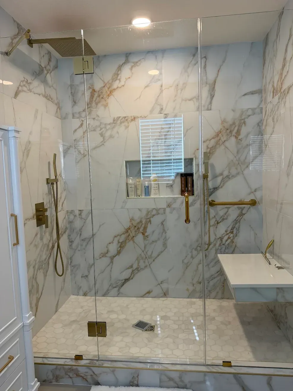 Modern bathroom with marble-look tiled shower, glass door, brass grab bars, bench, and handheld showerhead