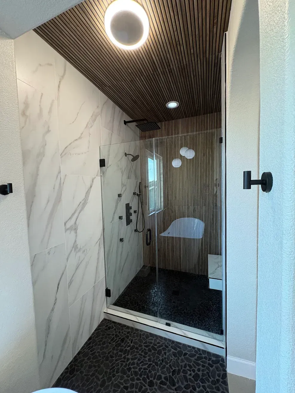 Narrow modern shower with marble walls, wood slat ceiling, and glass door in a bathroom
