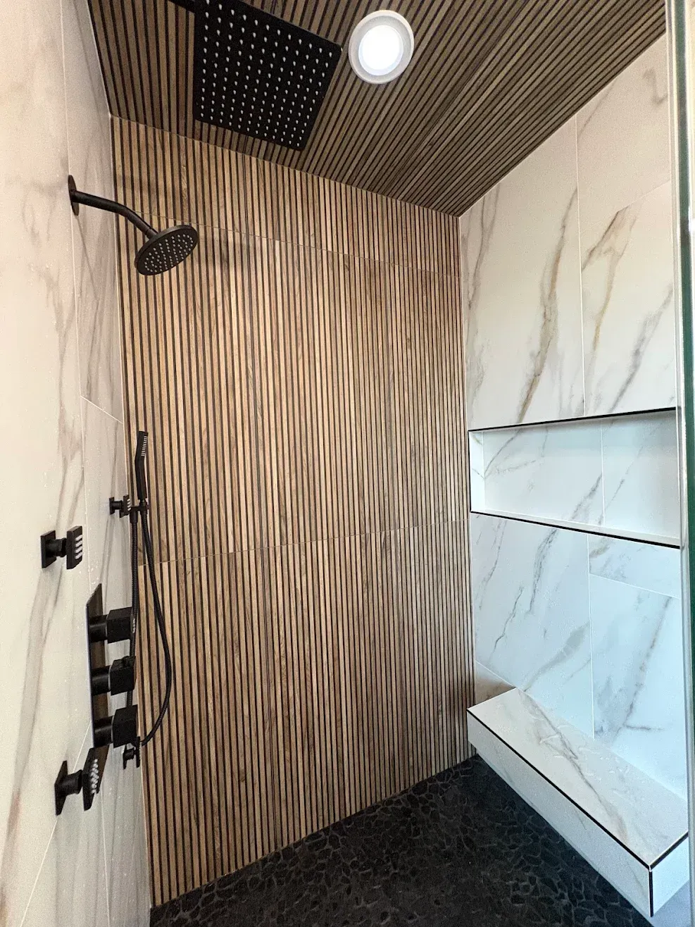 Modern shower with wood slat wall, marble tile, black fixtures, and a ceiling light