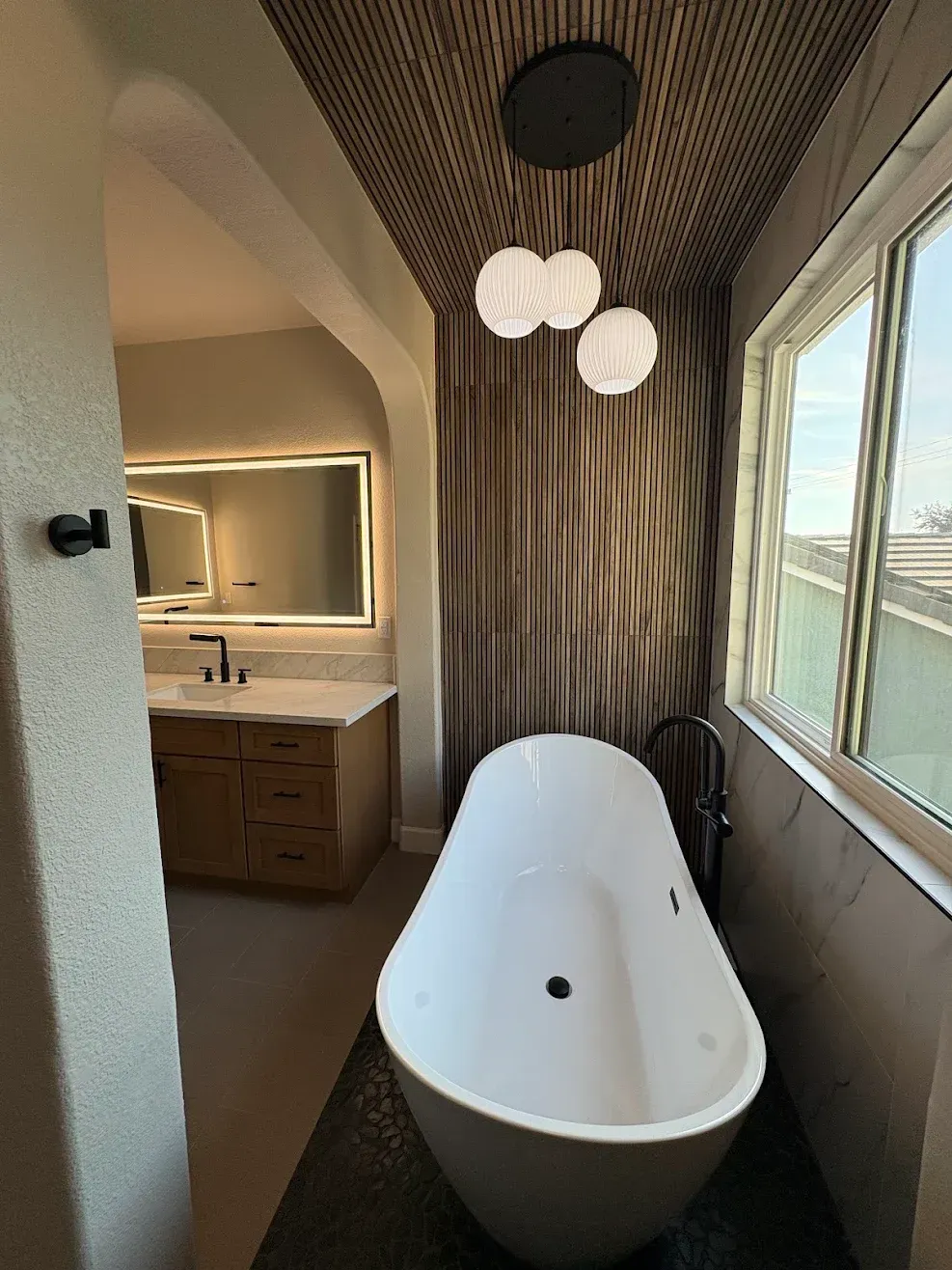 Modern bathroom with a freestanding white tub, wood-paneled wall, vanity, and large windows