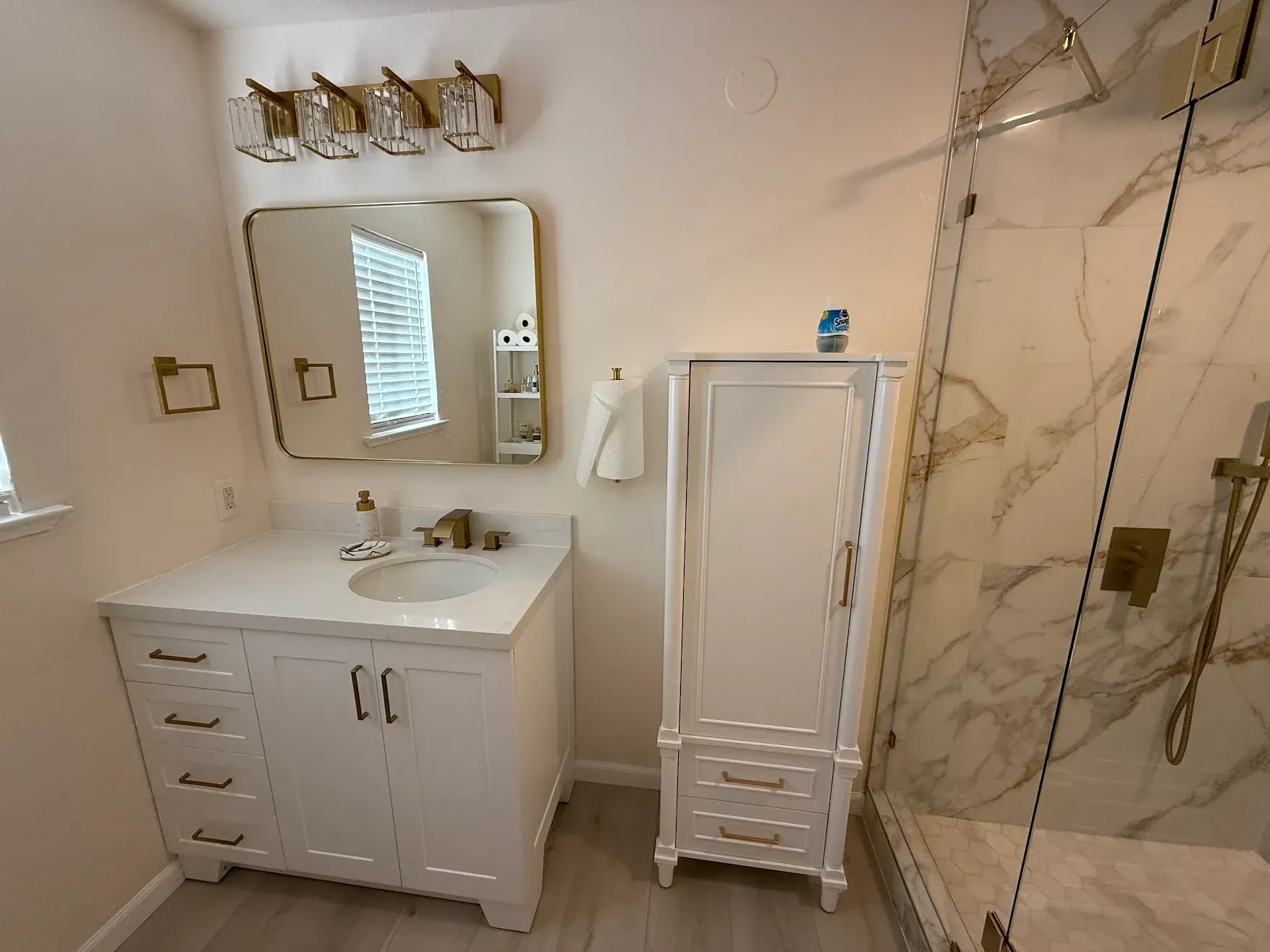White bathroom vanity with mirror and sconce lights beside a glass-enclosed marble shower.