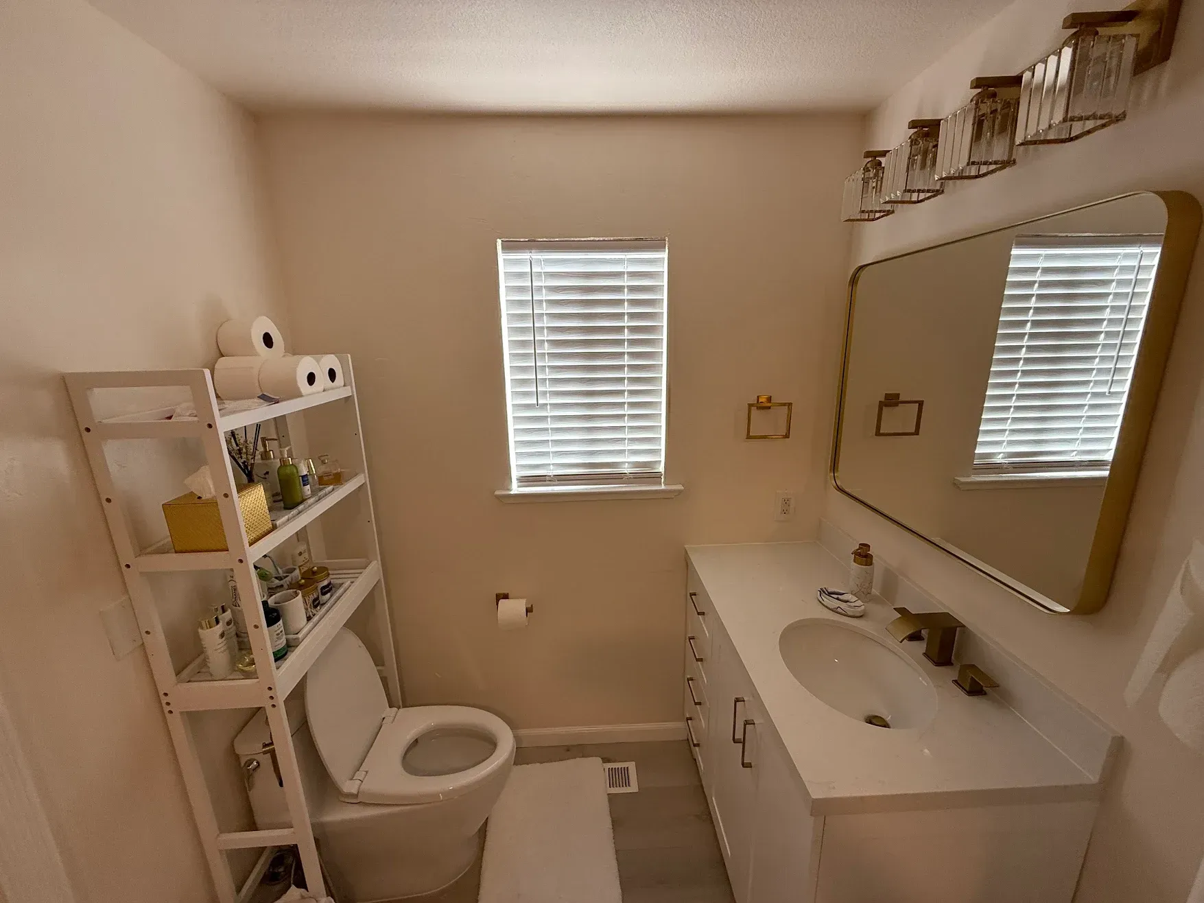 Small beige bathroom with toilet, sink, mirror, shelving, and window blinds.