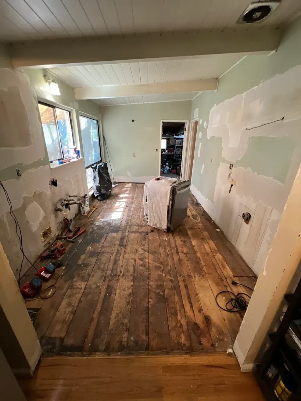 Narrow room under renovation with unfinished wooden floor, patchy walls, and tools along the sides.