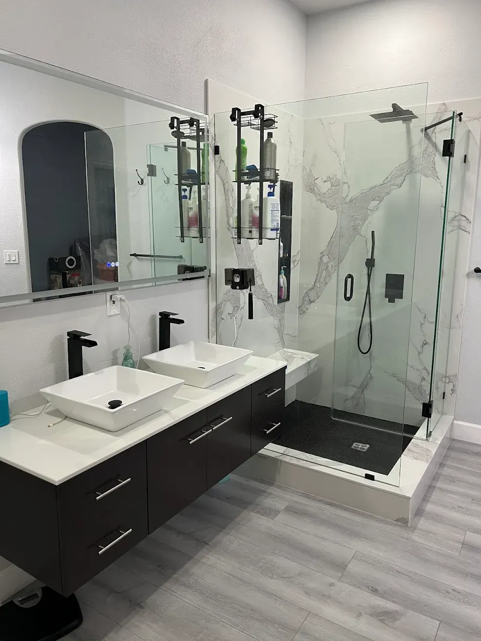 Modern bathroom with double sinks, large mirrors, and glass-enclosed shower; gray wood-look floor.