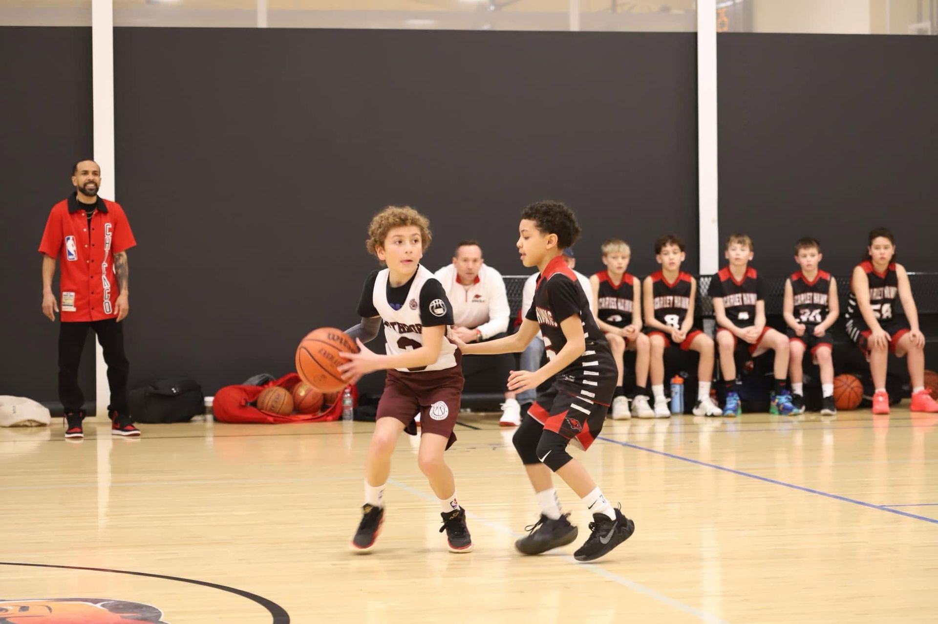 Youth basketball game in a gym, two players dribbling while teammates sit on the bench.