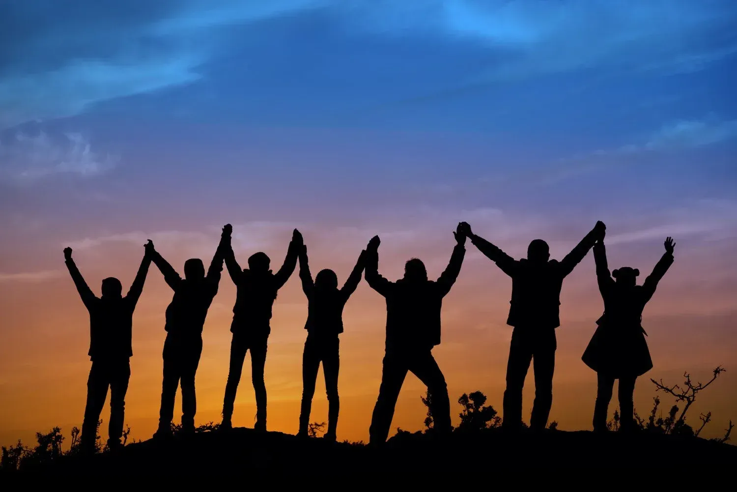 Un groupe de personnes se tenant la main, les bras levés, se détachant en silhouette sur une colline au coucher du soleil, avec un ciel coloré en arrière-plan.