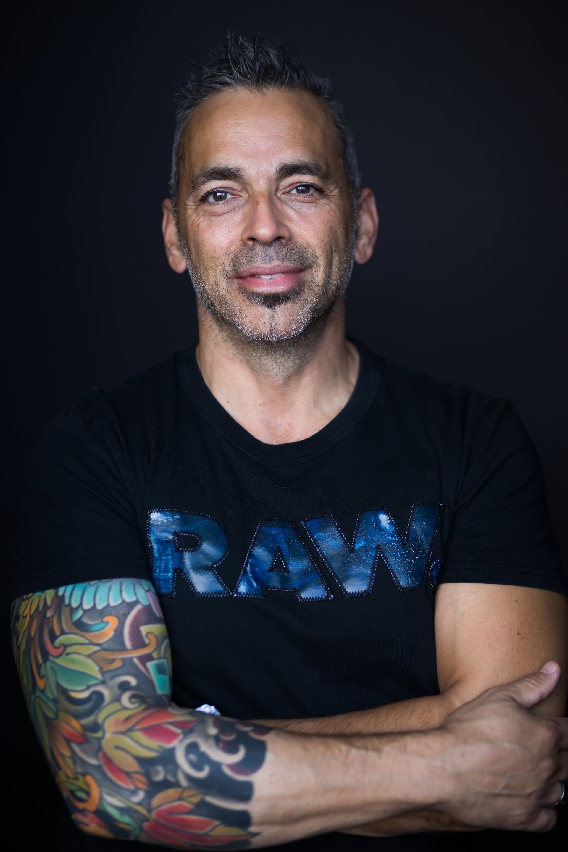 Un homme aux cheveux gris et tatoué, souriant, portant un t-shirt noir avec l'inscription « RAW », les bras croisés.