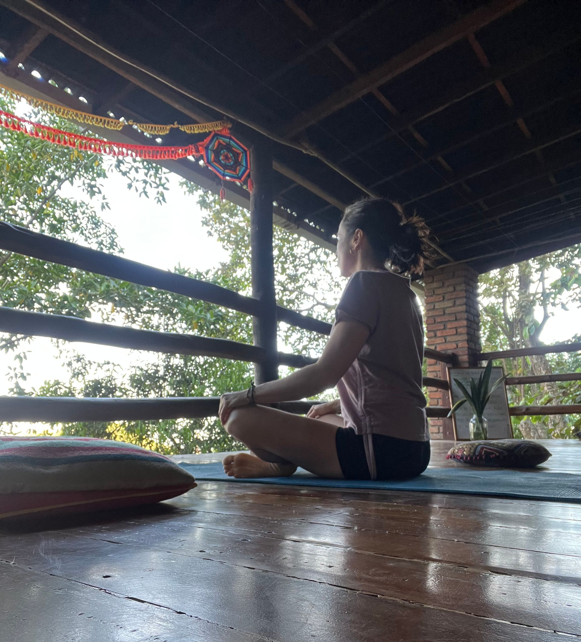 Femme assise en lotus sur un tapis de yoga, à l'extérieur, sous un abri, regardant au loin, pratiquant la méditation.