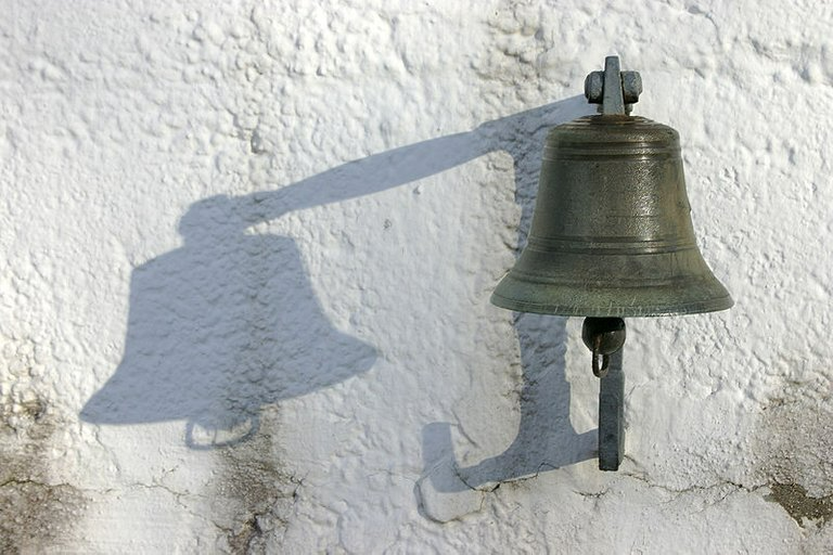 Cloche en bronze projetant une ombre sur un mur blanc et texturé.