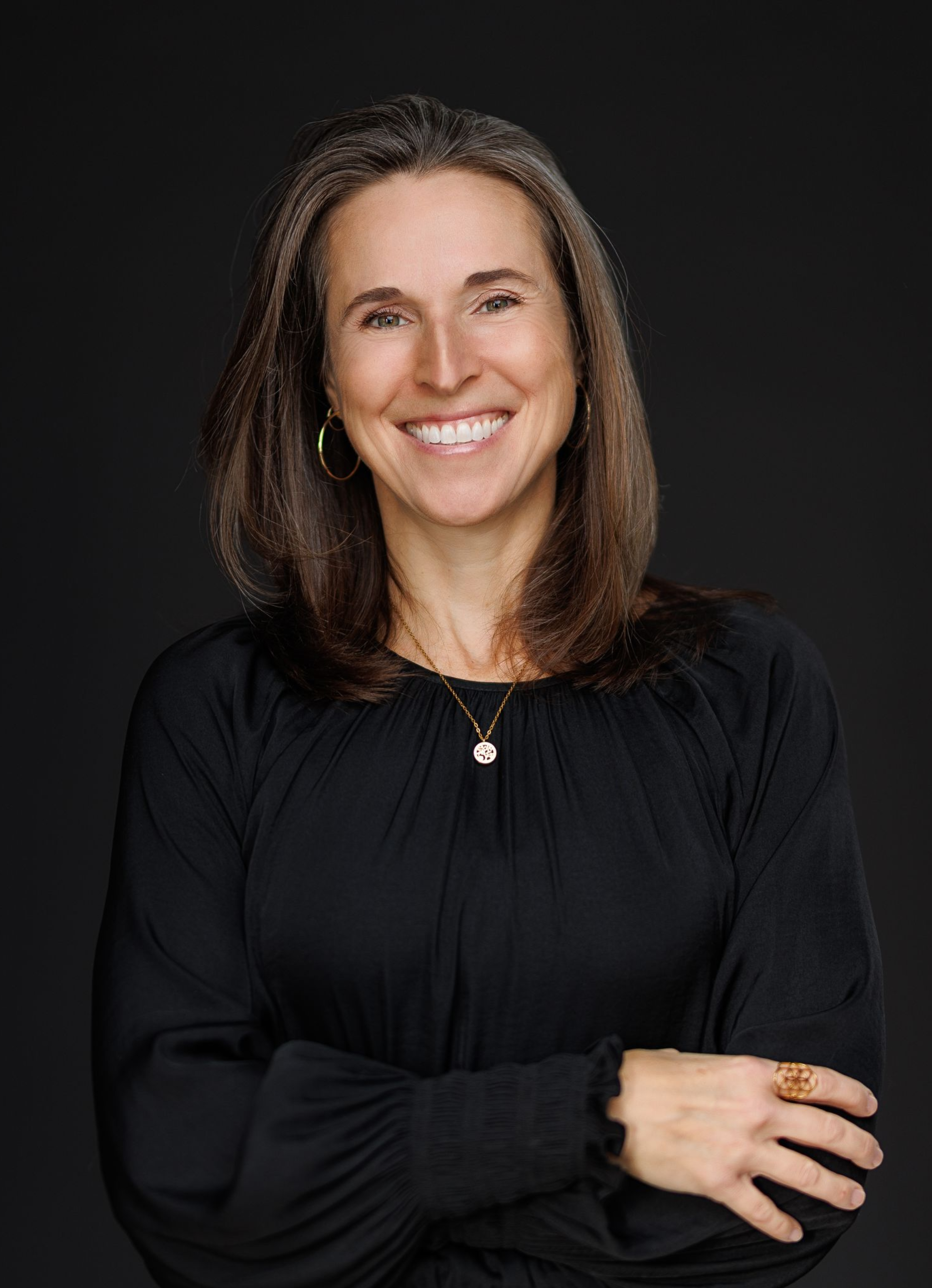 Femme aux longs cheveux bruns, souriante, vêtue d'un haut noir et de bijoux en or, sur fond sombre.