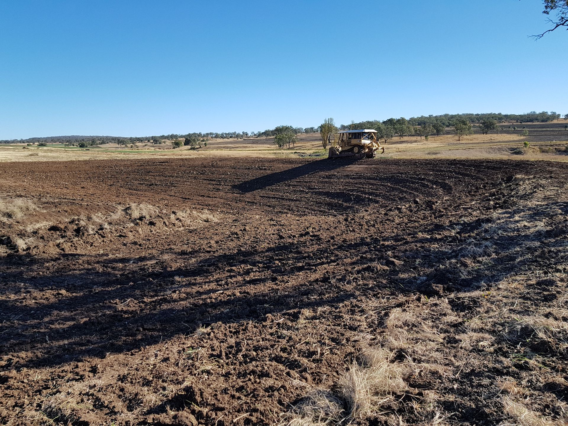 Wide Muddy Field With a Tractor in the Distance Under a Clear Blue Sky — Oakleigh Earthmoving in Chinchilla, QLD