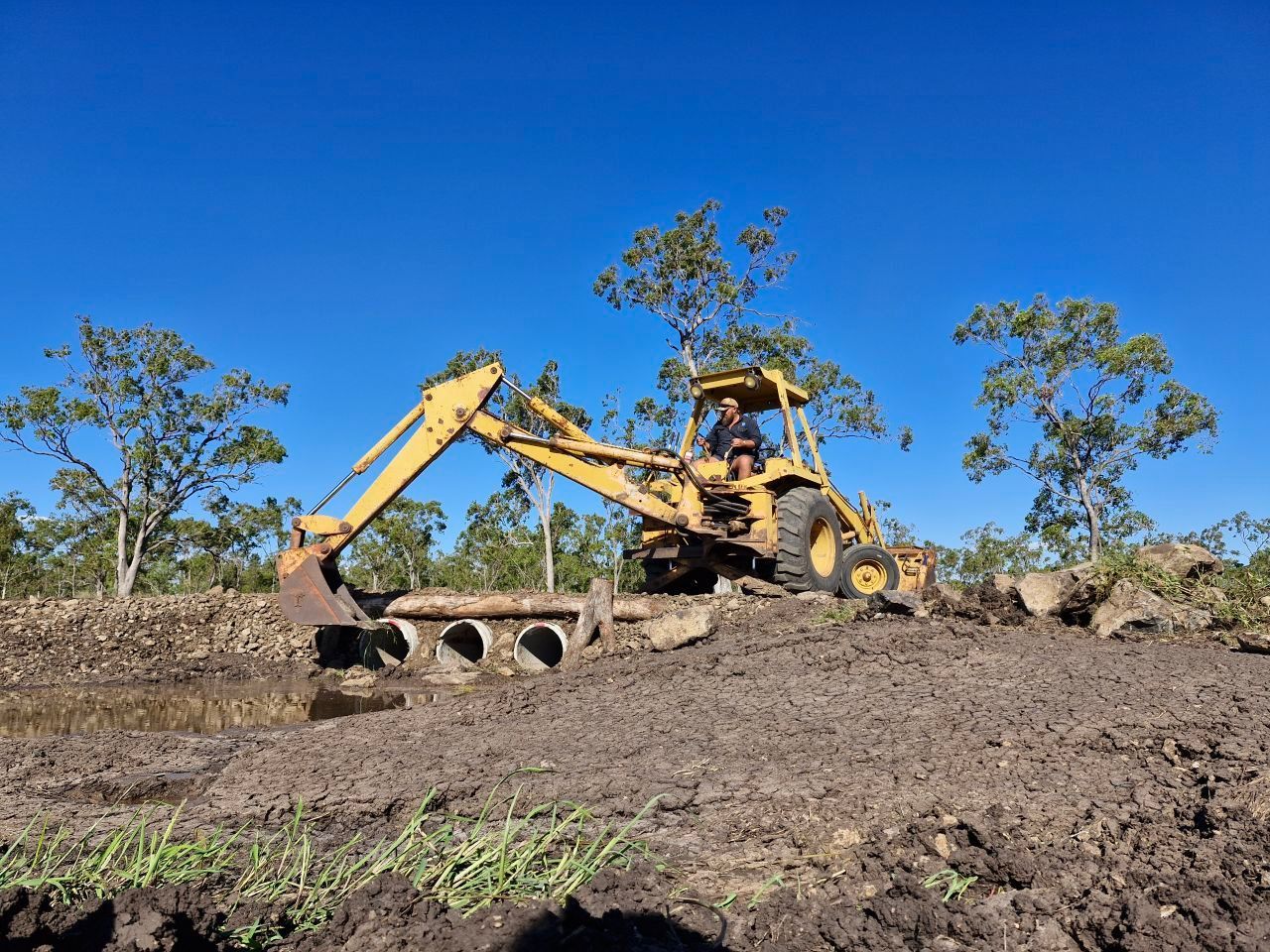 Yellow Excavator Digging Dirt in a Muddy Field Under a Clear Blue Sky — Oakleigh Earthmoving in Chinchilla, QLD