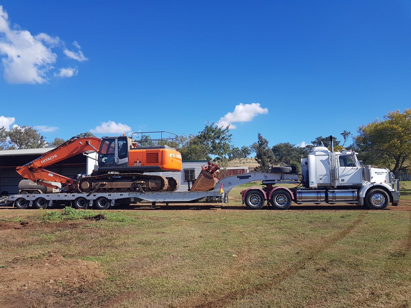 Flatbed Truck Hauling an Orange Excavator on a Rural Road Under a Blue Sky — Oakleigh Earthmoving in Warwick, QLD