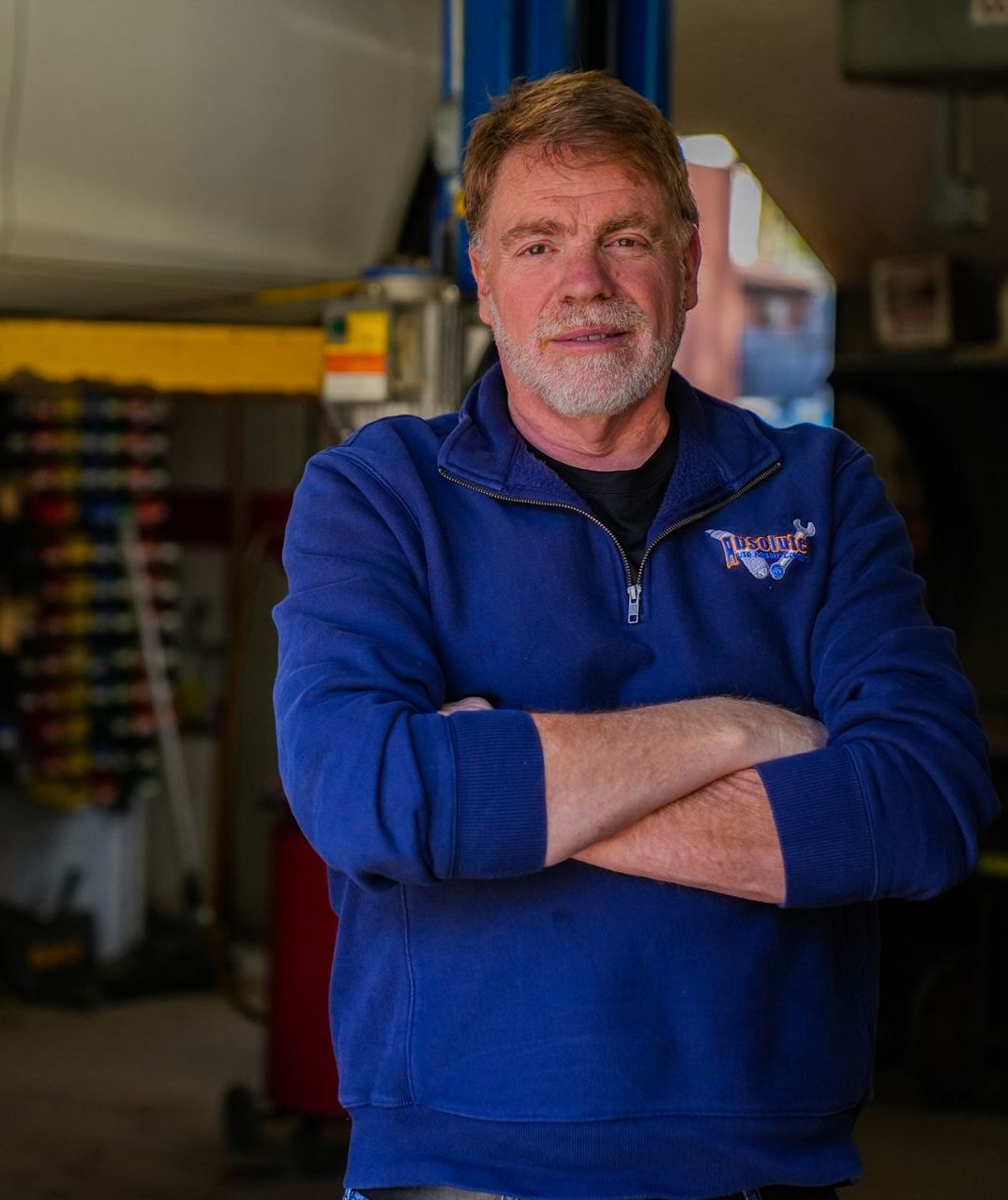 Man standing with arms crossed in a blue fleece inside a workshop, with tools and equipment in the background