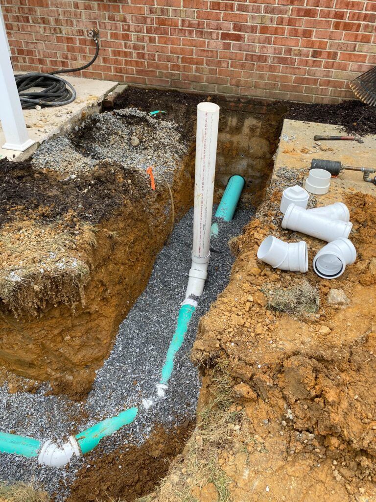 Excavated yard trench with white and green PVC pipes and fittings near a brick wall
