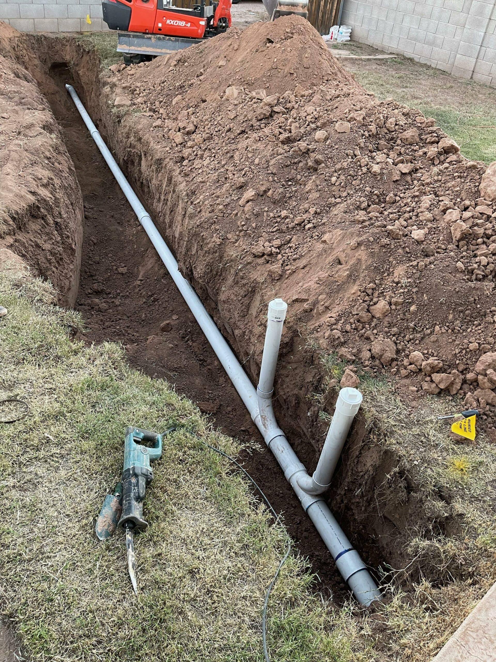 Trench with white PVC pipes being installed beside a dirt mound and an excavator on a grassy lot