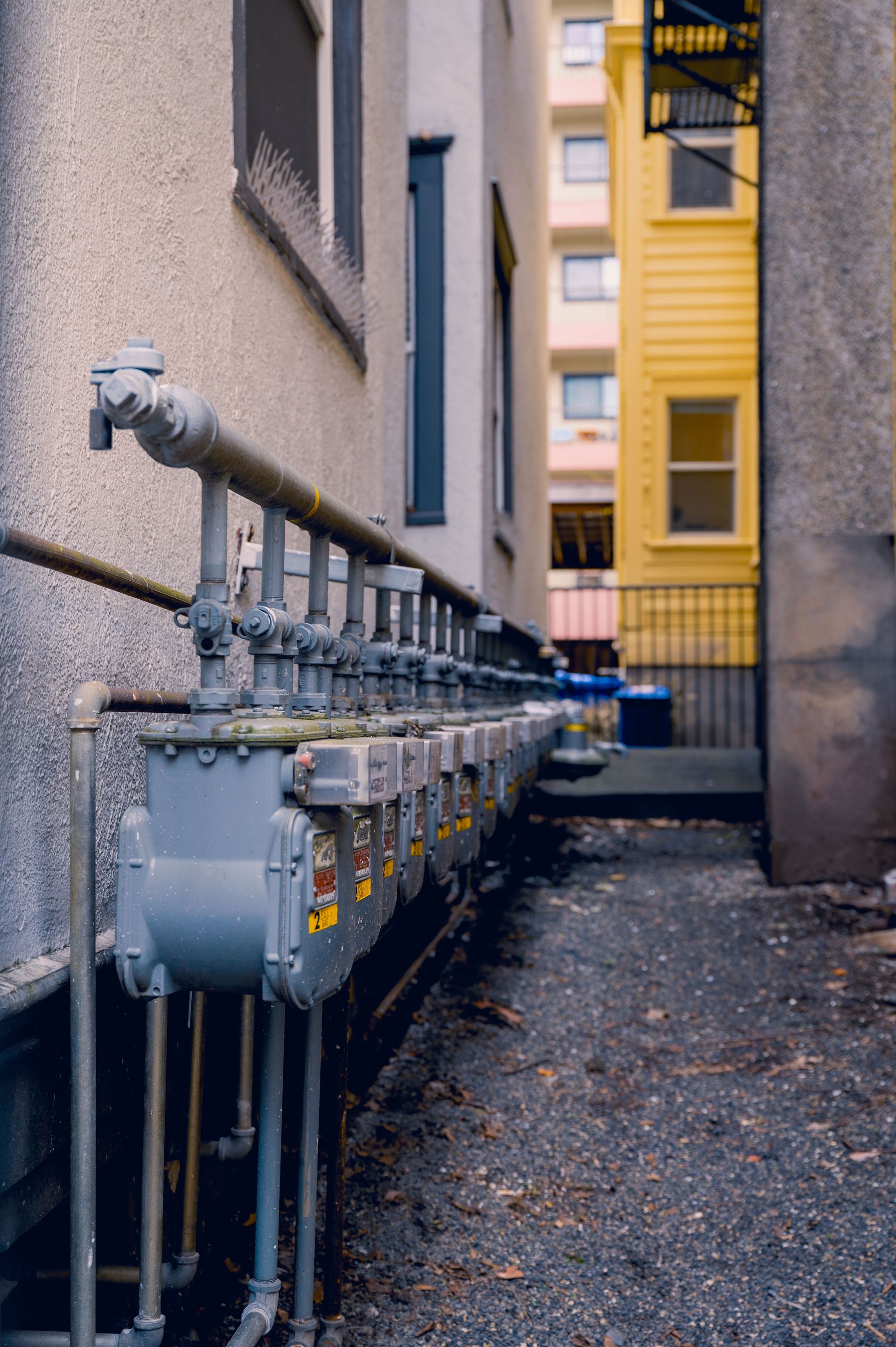 Row of gray utility meters and pipes beside a narrow alley, with a yellow building in the background
