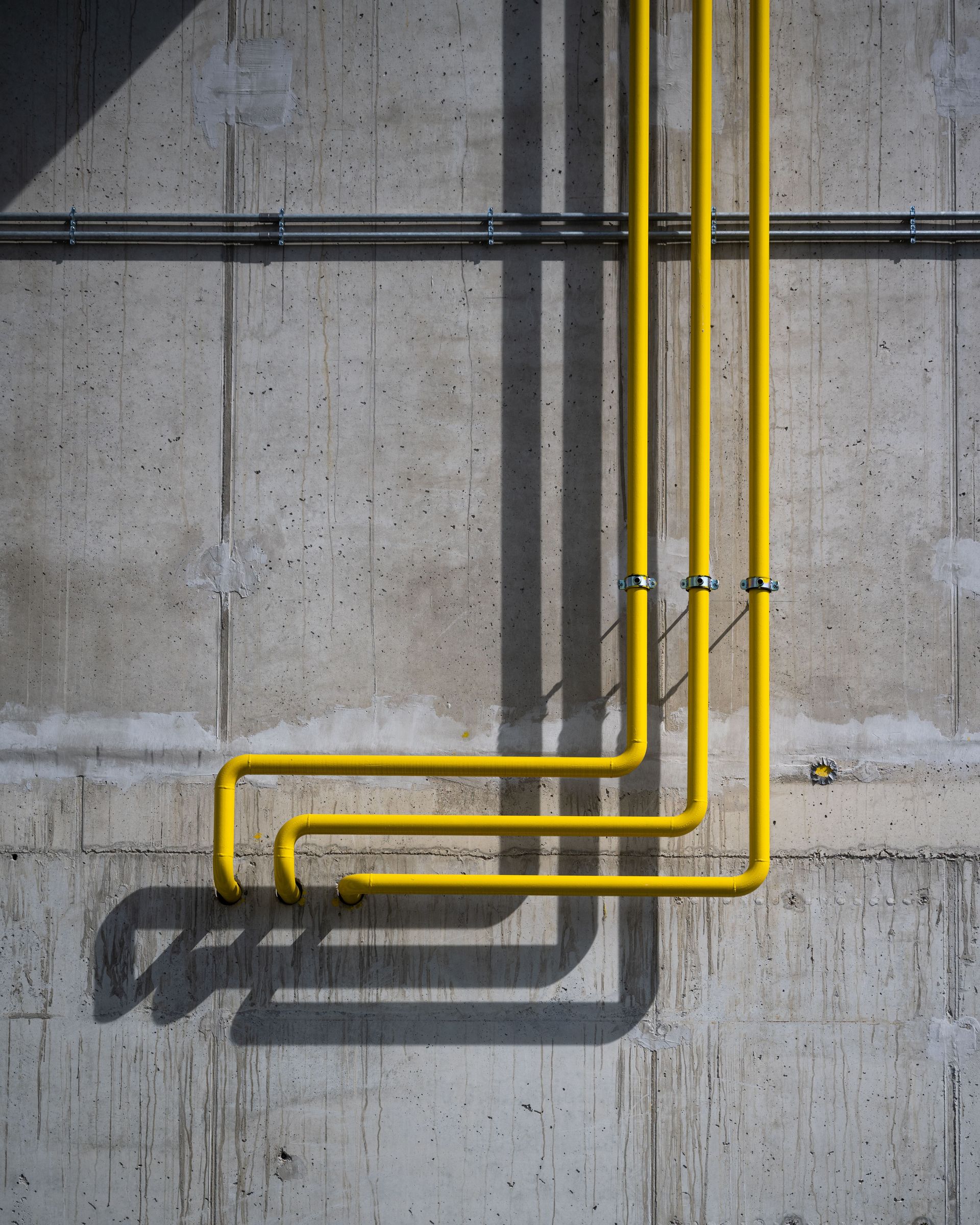 Yellow pipes running vertically and horizontally along a gray concrete wall, casting shadows.