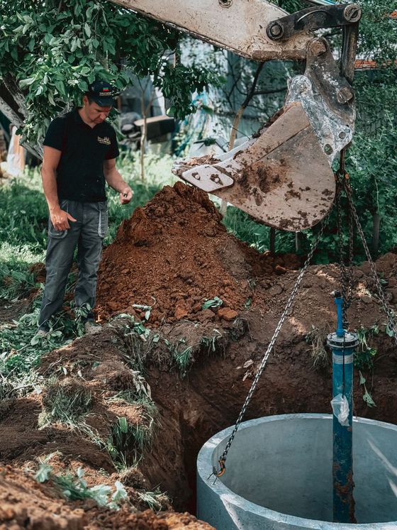 Excavator pouring soil into a concrete well pit as a worker stands nearby in a muddy outdoor area