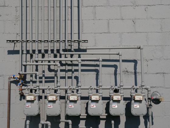 Row of electric meters and conduit pipes mounted on a gray concrete wall.