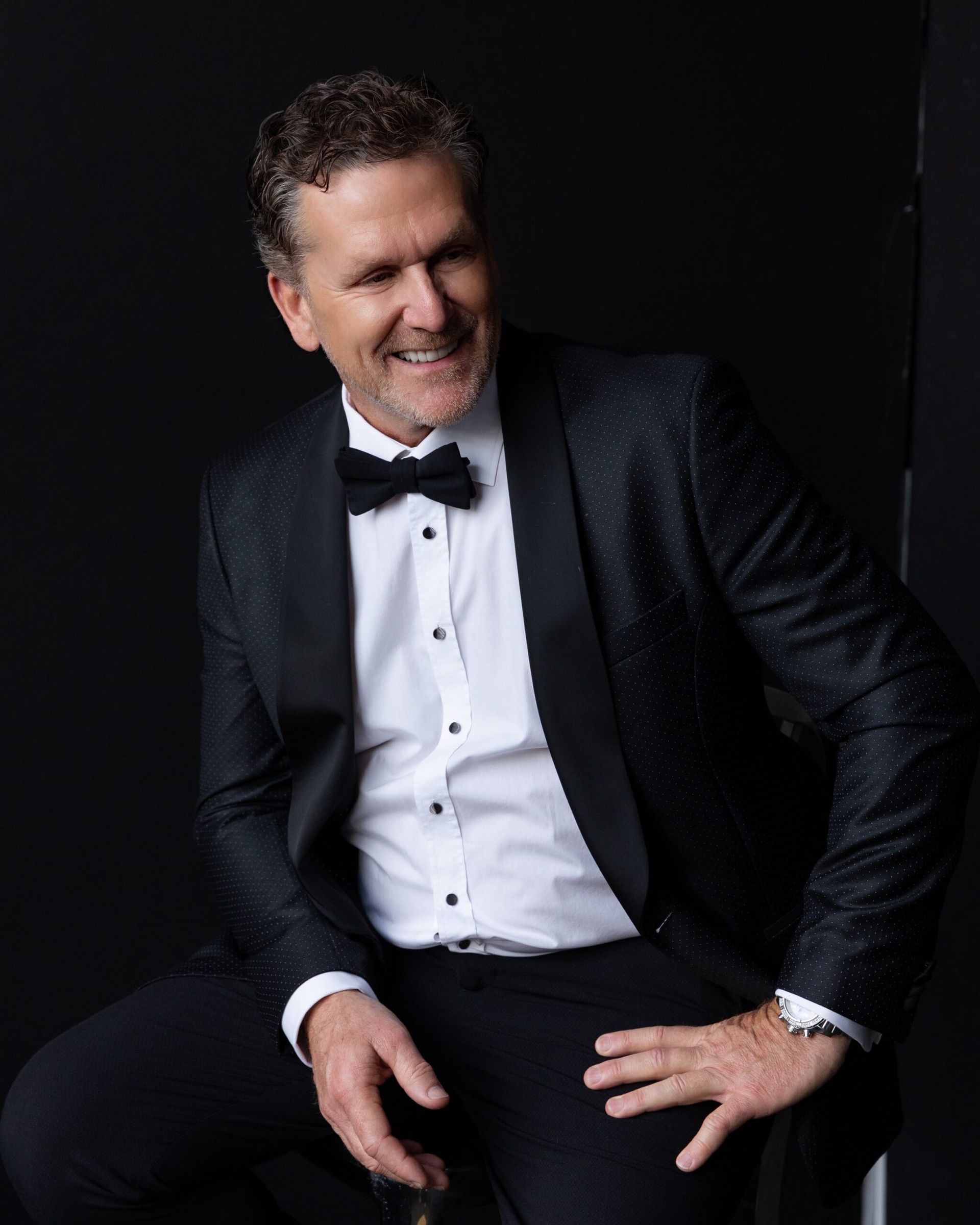 Dan Cooper in a tuxedo and bow tie is sitting on a chair