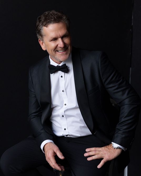 Dan Cooper in a tuxedo and bow tie is sitting on a chair