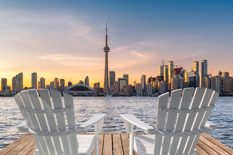 Two white adirondack chairs are sitting on a dock overlooking the city skyline at sunset.
