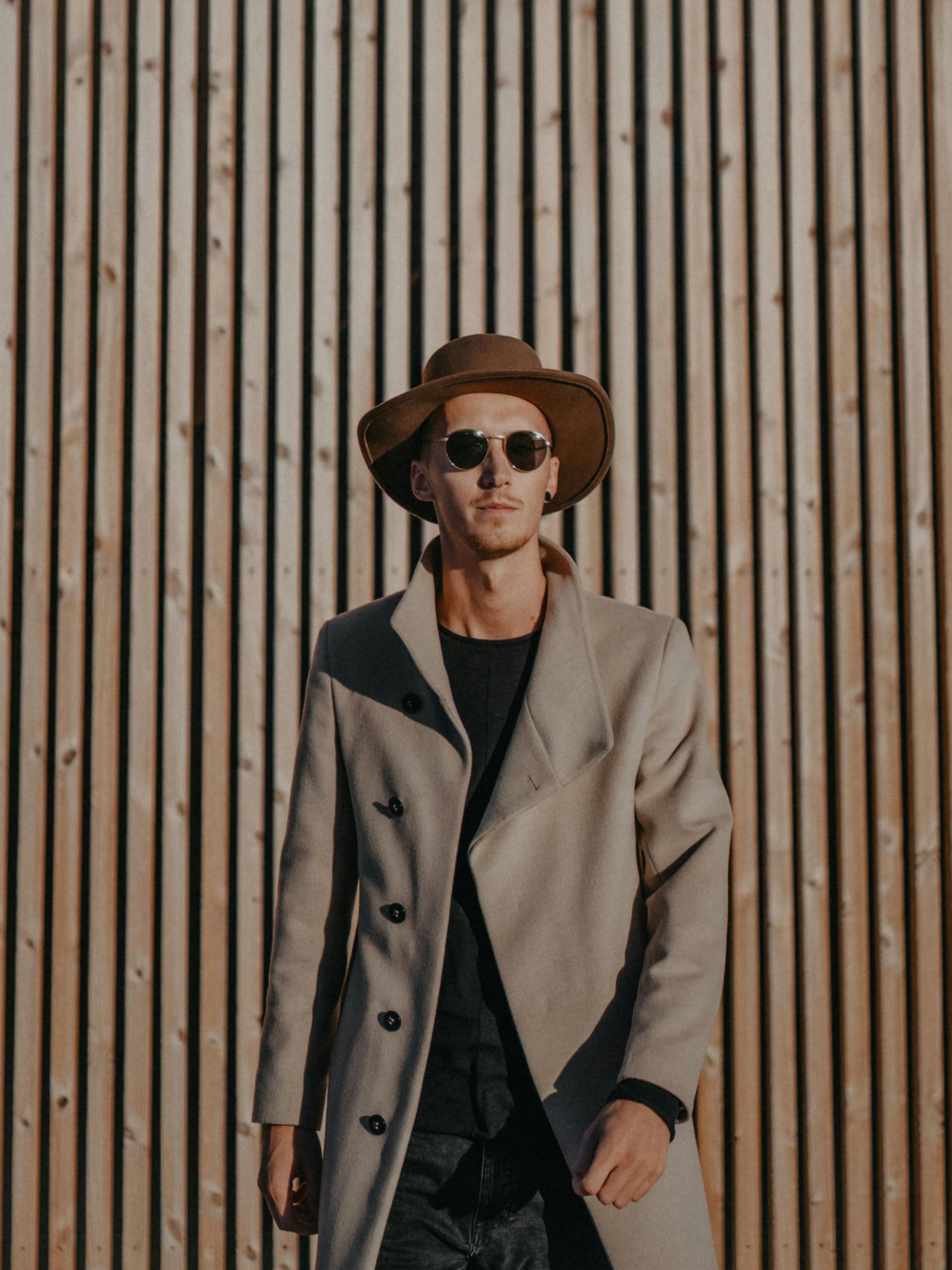 A man wearing a hat and sunglasses is standing in front of a wooden wall.